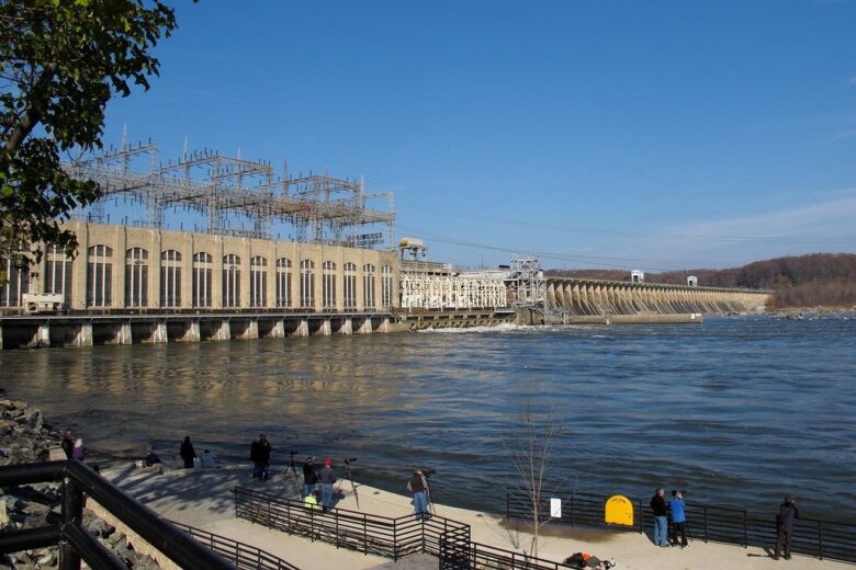Dam and power station beside a wide river with a few people on the waterfront under a clear blue sky.
