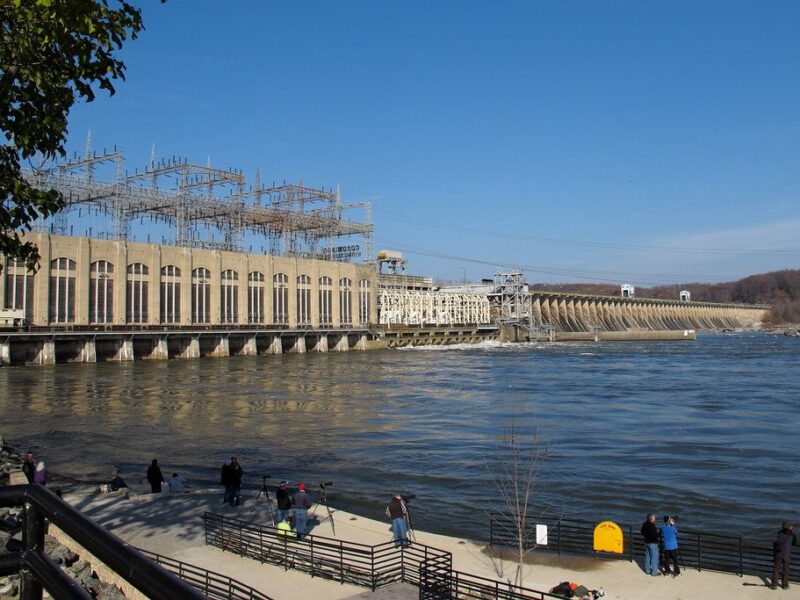 Dam and power station beside a wide river with a few people on the waterfront under a clear blue sky.
