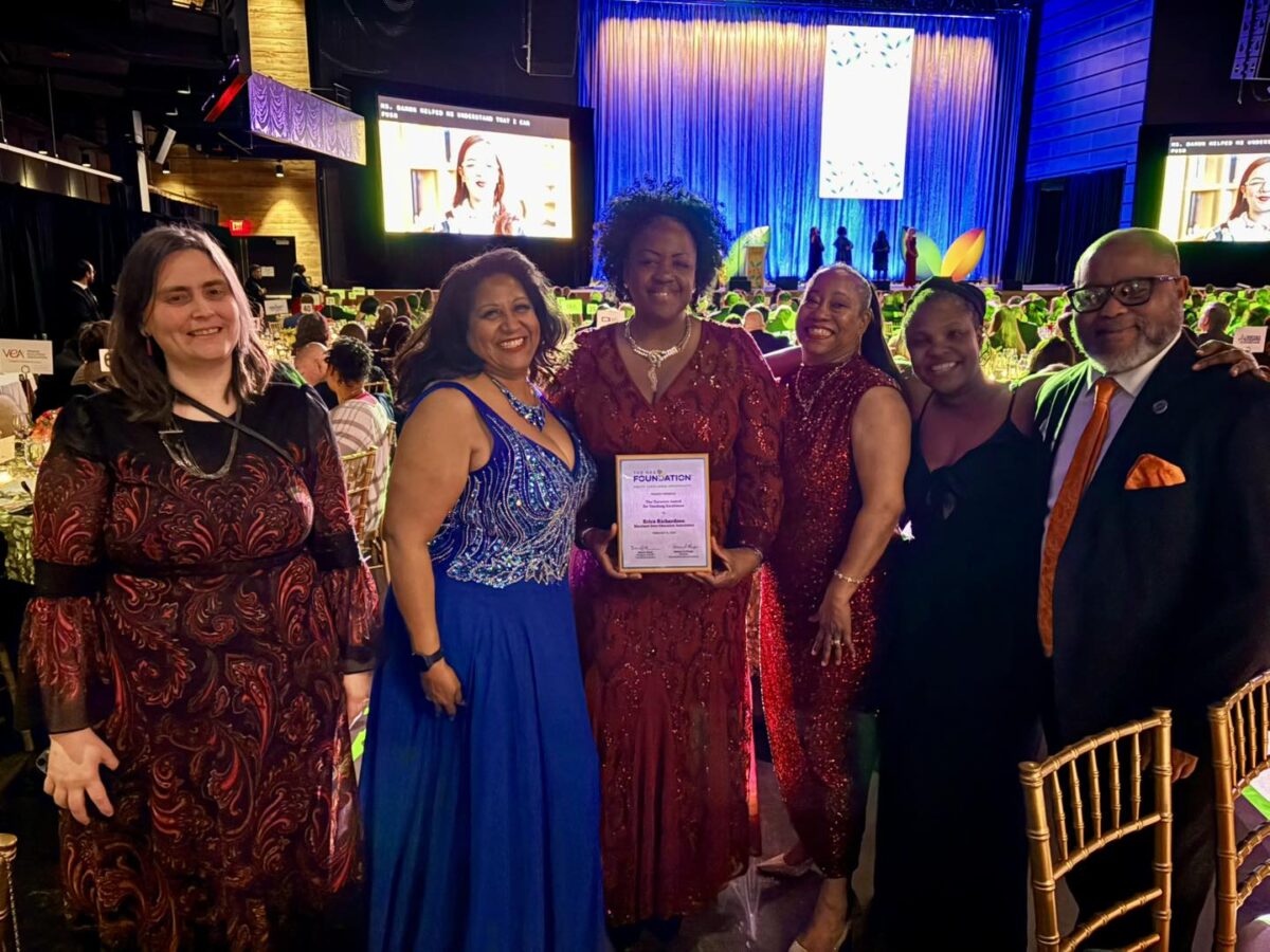 Group of six formally dressed people posing at an awards banquet holding a certificate on stage.