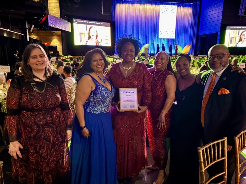 Group of six formally dressed people posing at an awards banquet holding a certificate on stage.