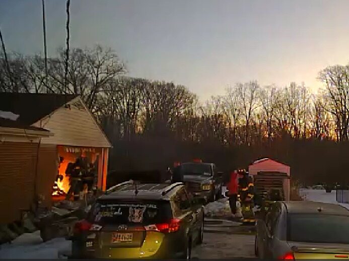 Firefighters work to control a garage fire beside parked cars at dusk.