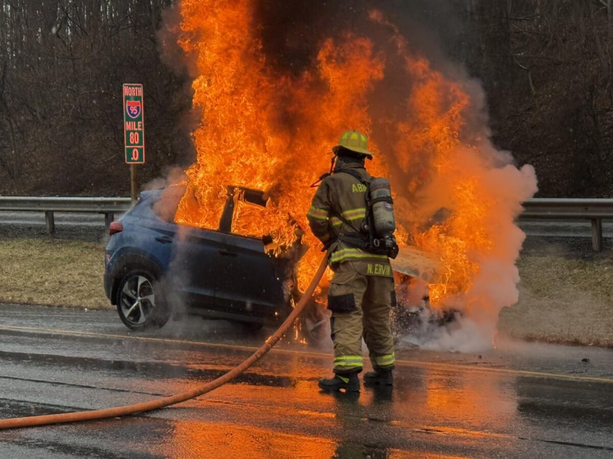 Firefighter battling a car engulfed in flames on a wet roadside