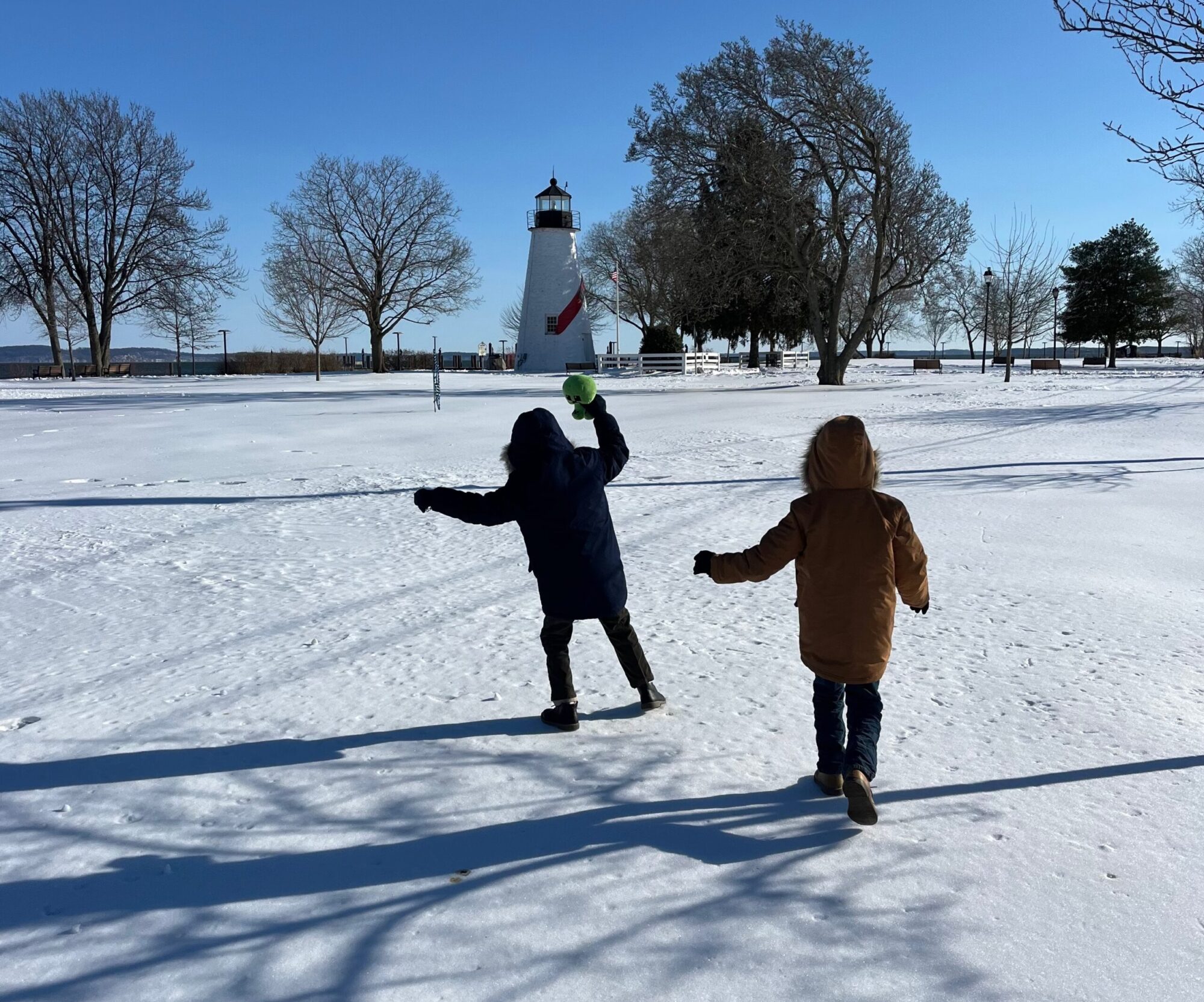 Two children run through a snowy park toward a lighthouse under a clear blue sky.