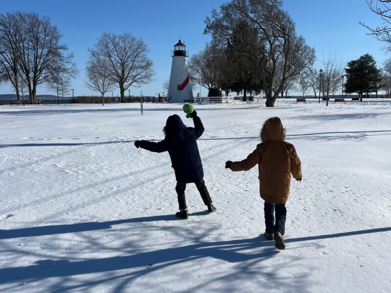 Two children run through a snowy park toward a lighthouse under a clear blue sky.