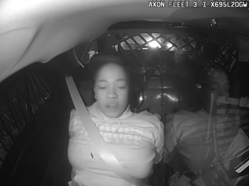 Woman asleep in the back of a police vehicle beside a reflected passenger, seen in black and white.