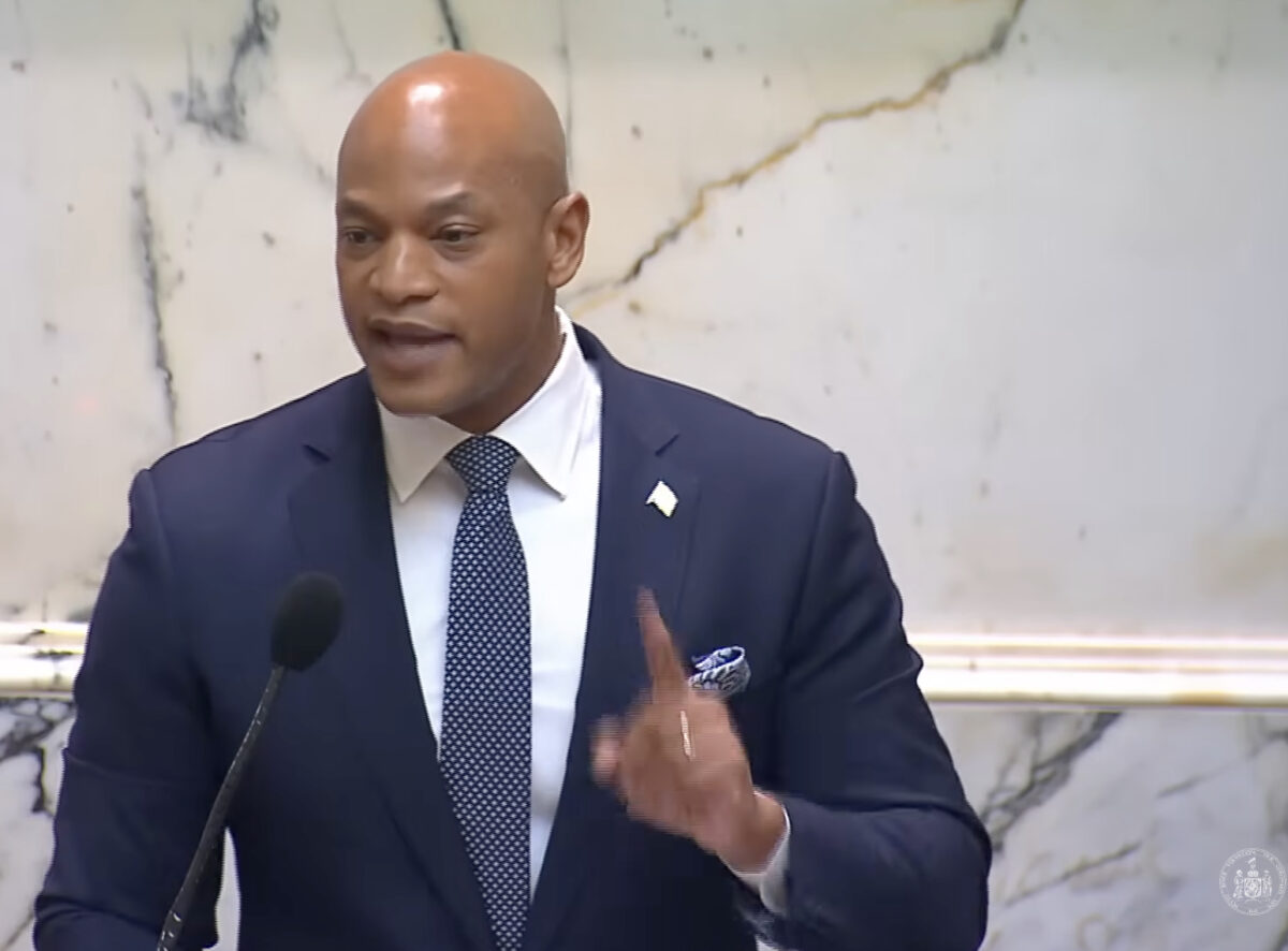 Man in a navy suit speaks at a podium with a microphone in a marble interior.