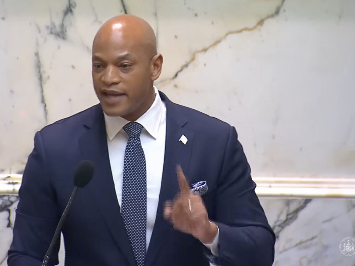 Man in a navy suit speaks at a podium with a microphone in a marble interior.