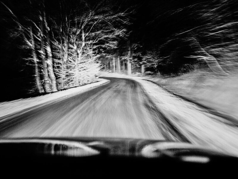 Blurred black-and-white view of a snowy road curving through a dark forest at night.
