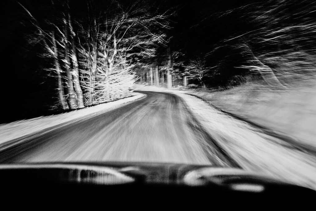 Blurred black-and-white view of a snowy road curving through a dark forest at night.