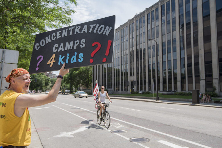 Protester holding a sign reading "Concentration Camps? 4 kids?!" on a city street.