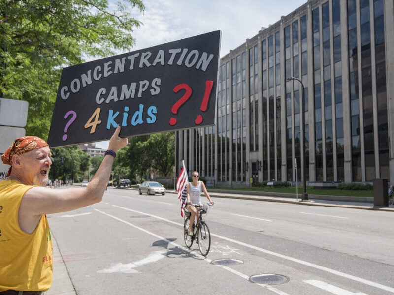 Protester holding a sign reading "Concentration Camps? 4 kids?!" on a city street.