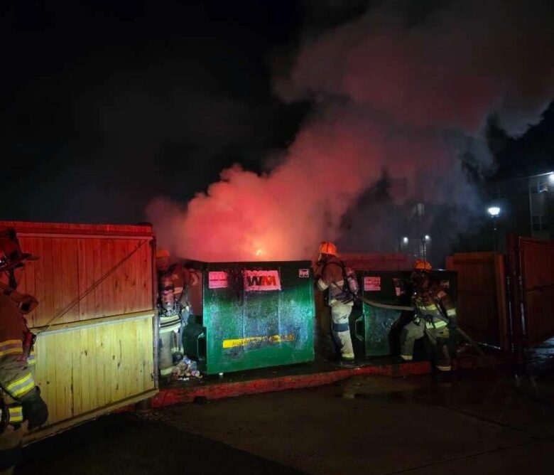 Firefighters battling a blaze beside dumpsters at night with thick smoke and red-orange glow.