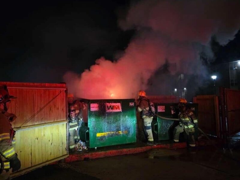 Firefighters battling a blaze beside dumpsters at night with thick smoke and red-orange glow.