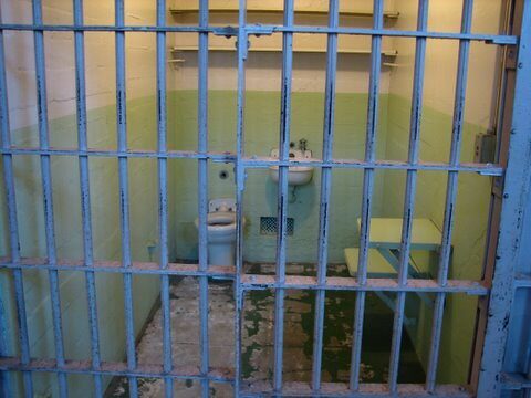 Empty prison cell with barred door, sink, toilet, and peeling green walls.