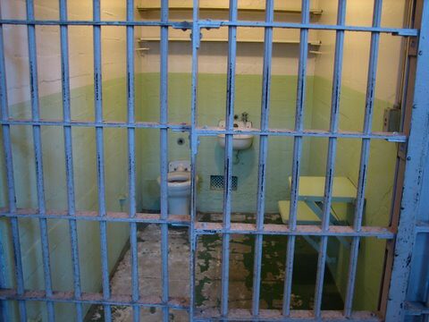Empty prison cell with barred door, sink, toilet, and peeling green walls.