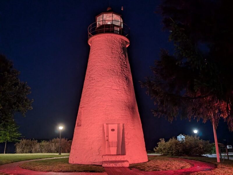 Pink-lit lighthouse at night with trees and a dark blue sky