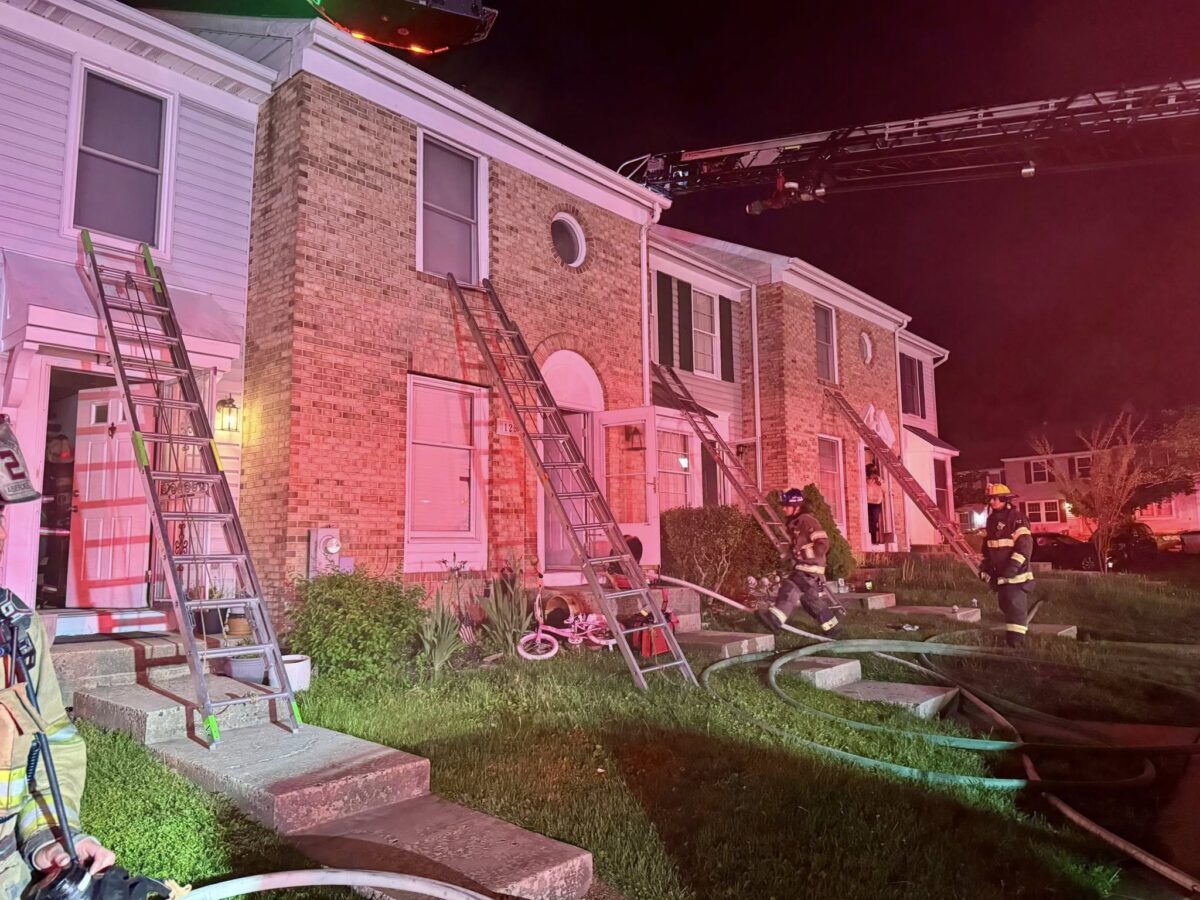 Firefighters work around ladders and hoses outside a brick townhouse at night under pink emergency lights.
