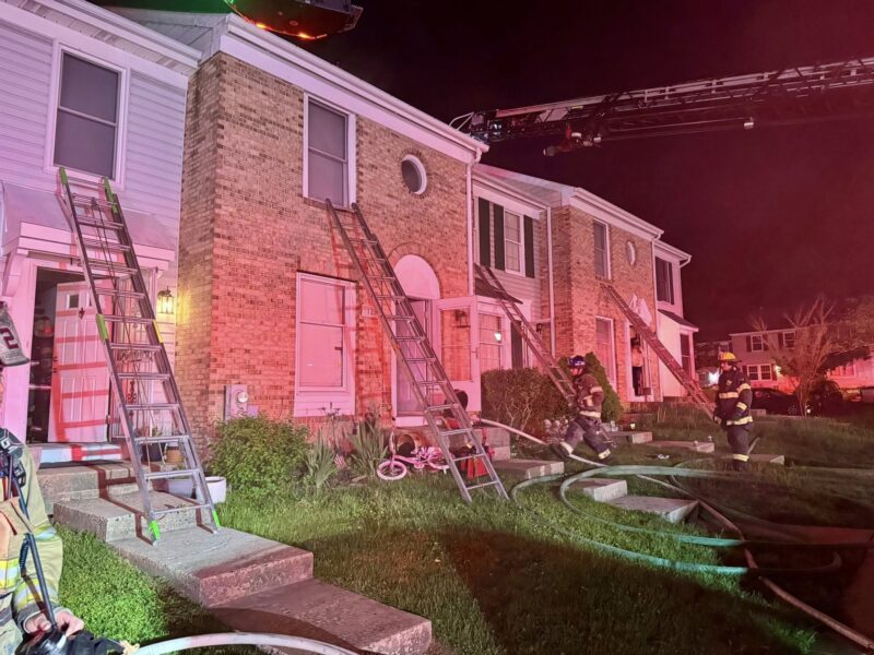 Firefighters work around ladders and hoses outside a brick townhouse at night under pink emergency lights.