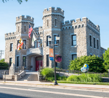 Historic stone building with castle-like towers, flags, and a front entrance by the street.