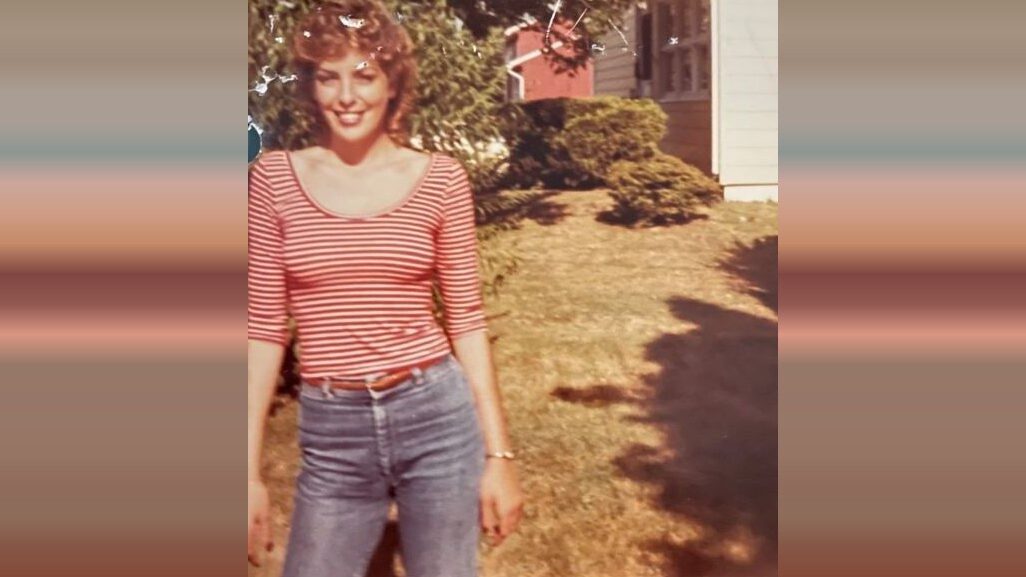 Smiling woman in a red striped top and jeans stands in a sunny yard beside a house.