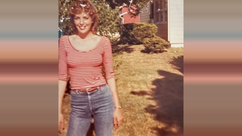 Smiling woman in a red striped top and jeans stands in a sunny yard beside a house.