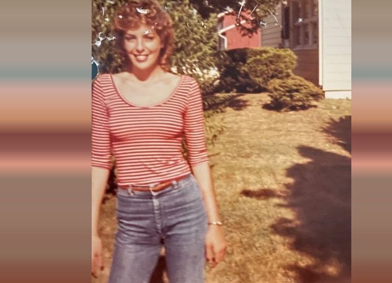 Smiling woman in a red striped top and jeans stands in a sunny yard beside a house.