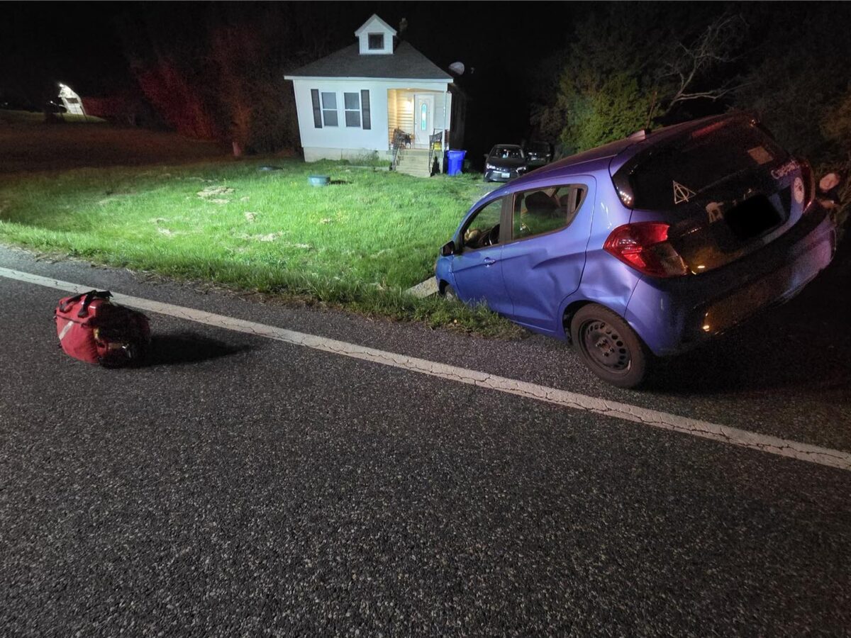 Blue car crashed off the road beside a small house at night with a red bag on the pavement.