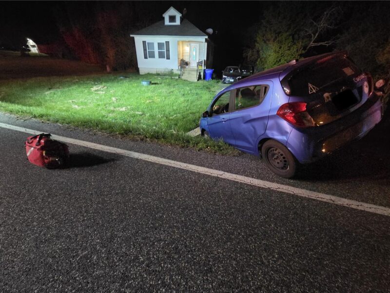 Blue car crashed off the road beside a small house at night with a red bag on the pavement.