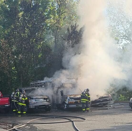 Firefighters battle a vehicle blaze with thick smoke rising beside parked trucks in a wooded area.