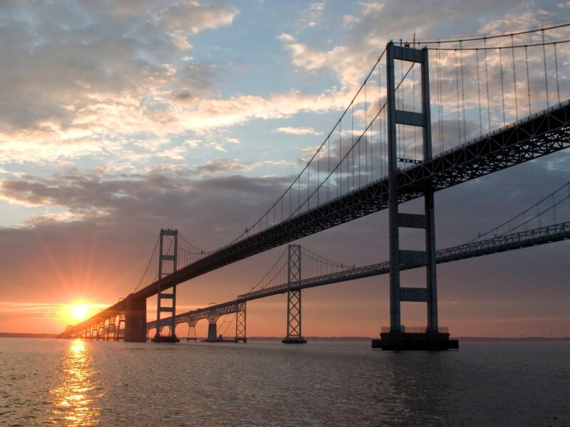 Suspension bridge at sunset over calm water with glowing sky and reflections