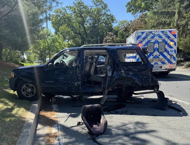 Damaged black SUV with a detached car seat beside it and an ambulance parked behind on a sunny roadside.