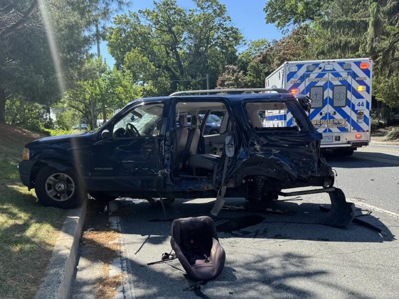 Damaged black SUV with a detached car seat beside it and an ambulance parked behind on a sunny roadside.