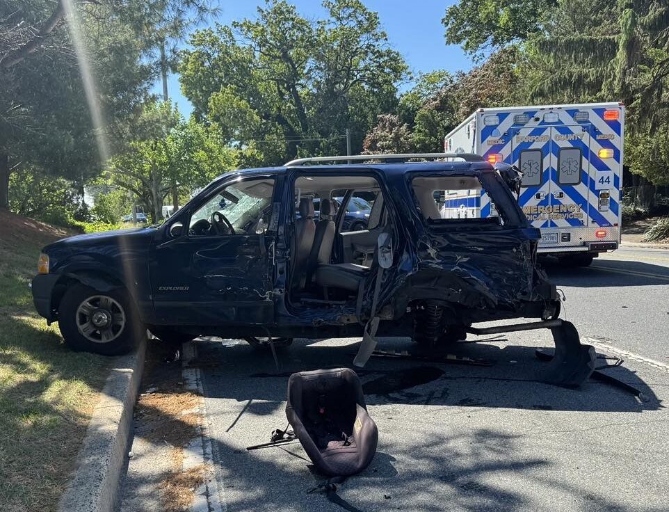 Damaged black SUV with a detached car seat beside it and an ambulance parked behind on a sunny roadside.