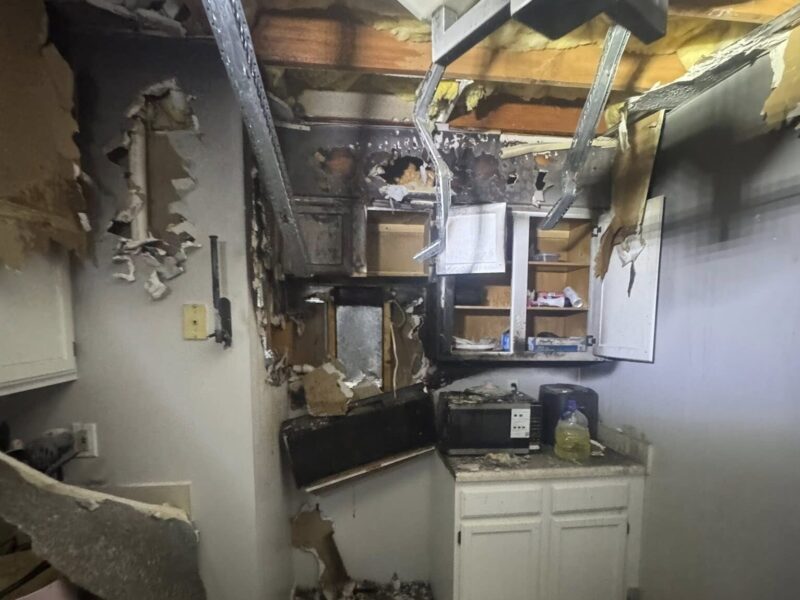 Severely fire-damaged kitchen with charred cabinets, debris, and exposed ceiling beams.