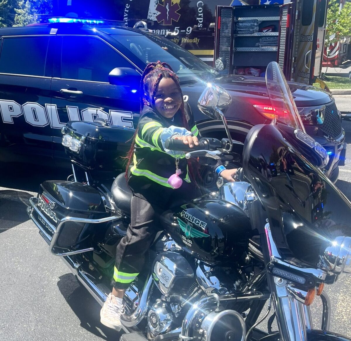 Child on a police motorcycle in front of a police SUV with flashing lights.