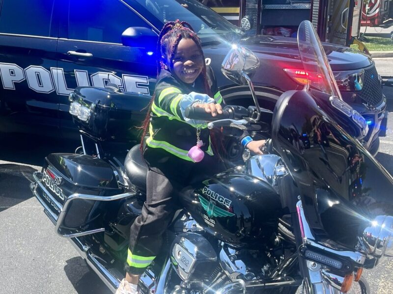 Child on a police motorcycle in front of a police SUV with flashing lights.