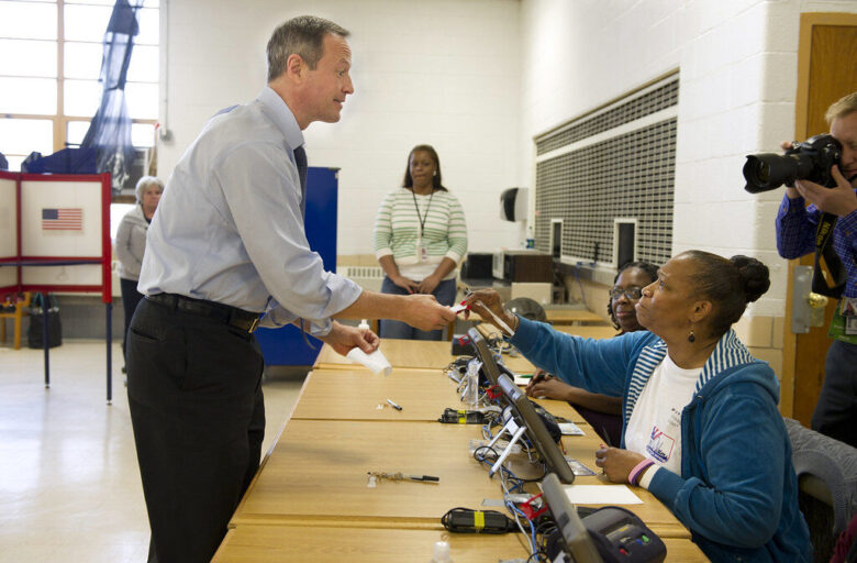 Man greeting a seated woman at a voting check-in table in a community polling place.