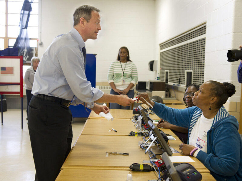 Man greeting a seated woman at a voting check-in table in a community polling place.