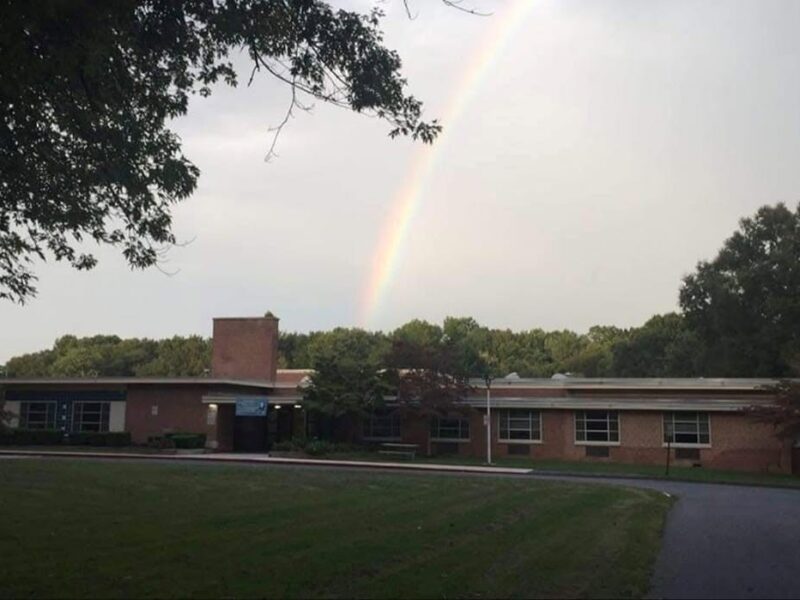 Rainbow above a low brick school building with trees and a grassy lawn.