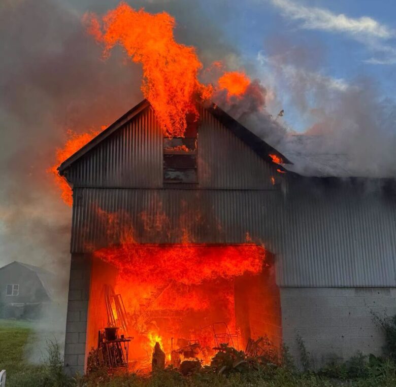 Large barn engulfed in flames and smoke at dusk with fire pouring from the roof and doorway.