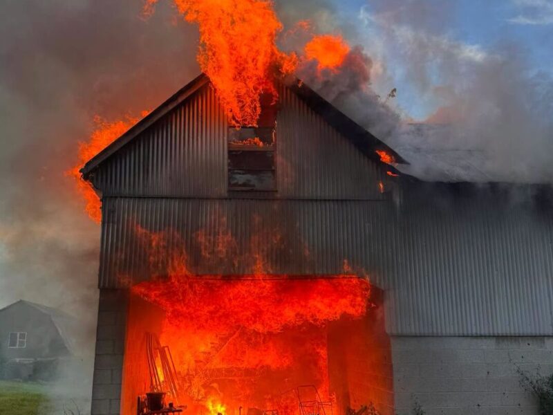 Large barn engulfed in flames and smoke at dusk with fire pouring from the roof and doorway.