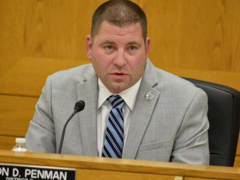 Man in a gray suit speaking at a microphone in a wood-paneled meeting room.