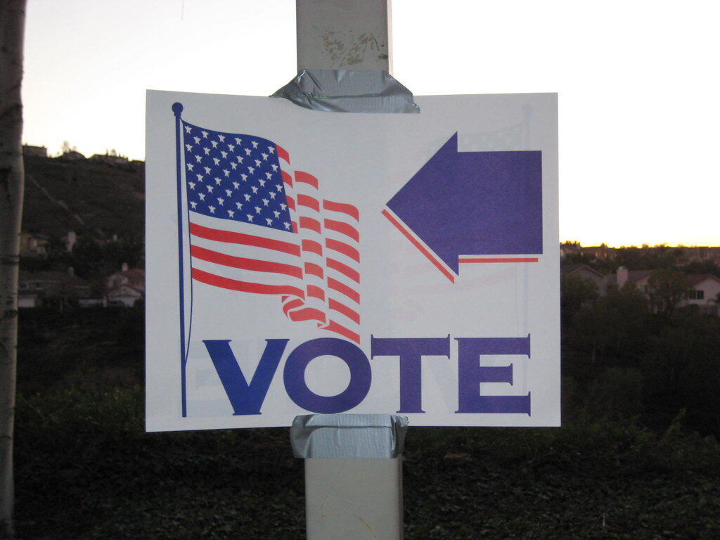Handmade voting sign with an American flag, large arrow, and the word VOTE taped to a pole at dusk.