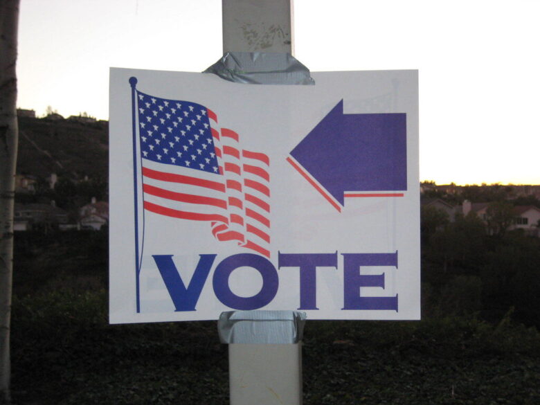 Handmade voting sign with an American flag, large arrow, and the word VOTE taped to a pole at dusk.