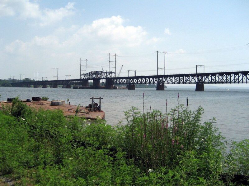 Long steel bridge spans a calm river with green shoreline in the foreground under a partly cloudy sky.
