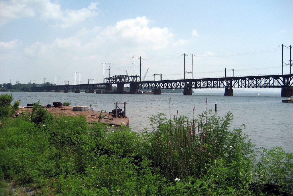 Long steel bridge spans a calm river with green shoreline in the foreground under a partly cloudy sky.