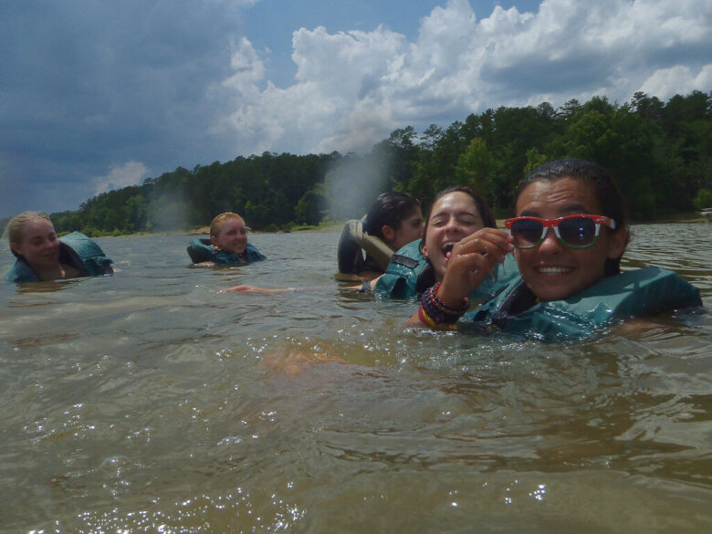 Group of friends floating in a lake with life jackets, smiling near a forested shoreline.