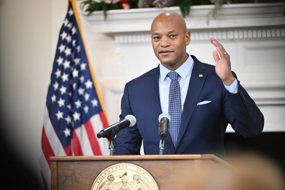 Man in a suit speaks at a podium with microphones beside a U.S. flag.