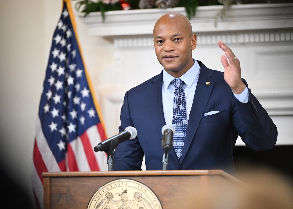 Man in a suit speaks at a podium with microphones beside a U.S. flag.