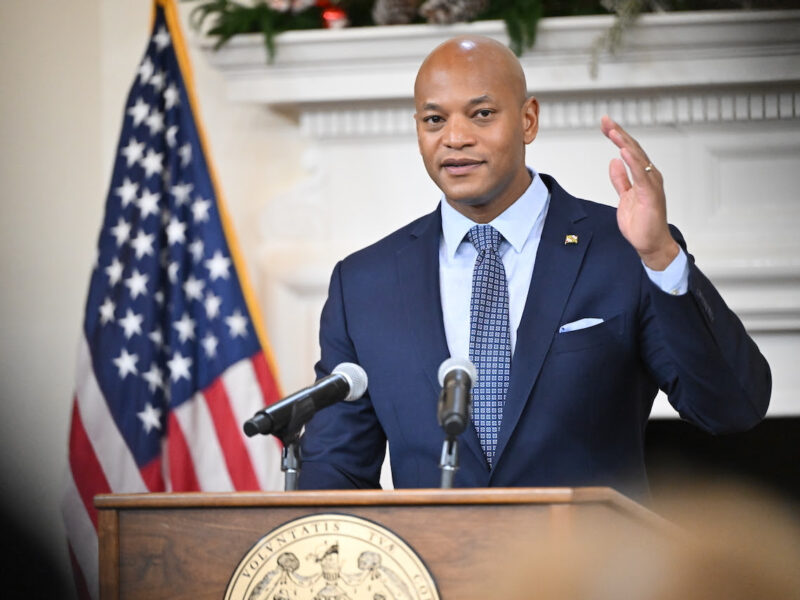 Man in a suit speaks at a podium with microphones beside a U.S. flag.